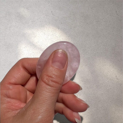 A hand holding a smooth, oval-shaped light pink worry stone made of rose quartz, against a textured light grey background.