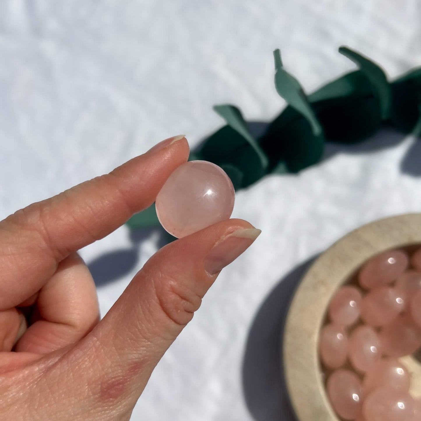 A small pink rose quartz crystal tumble stone held between finger and thumb with a bowl of crystals in the background