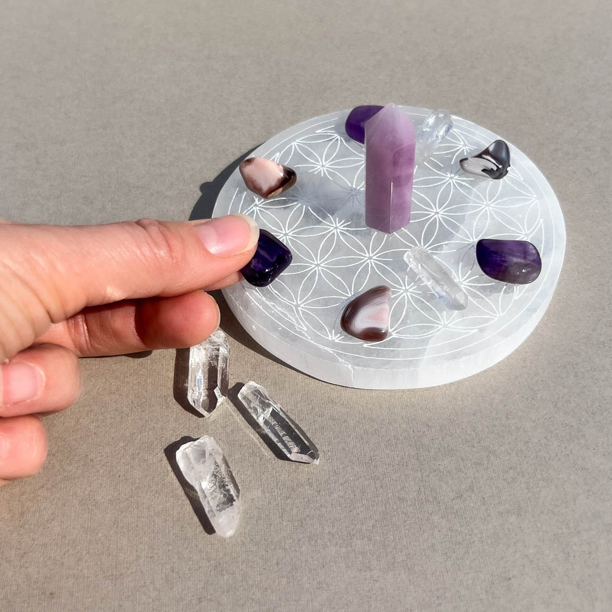 A hand holds a small amethyst crystal placing it in a grid formation on a selenite plate with a geometric flower of life pattern, with several loose clear quartz points on a neutral grey surface.
