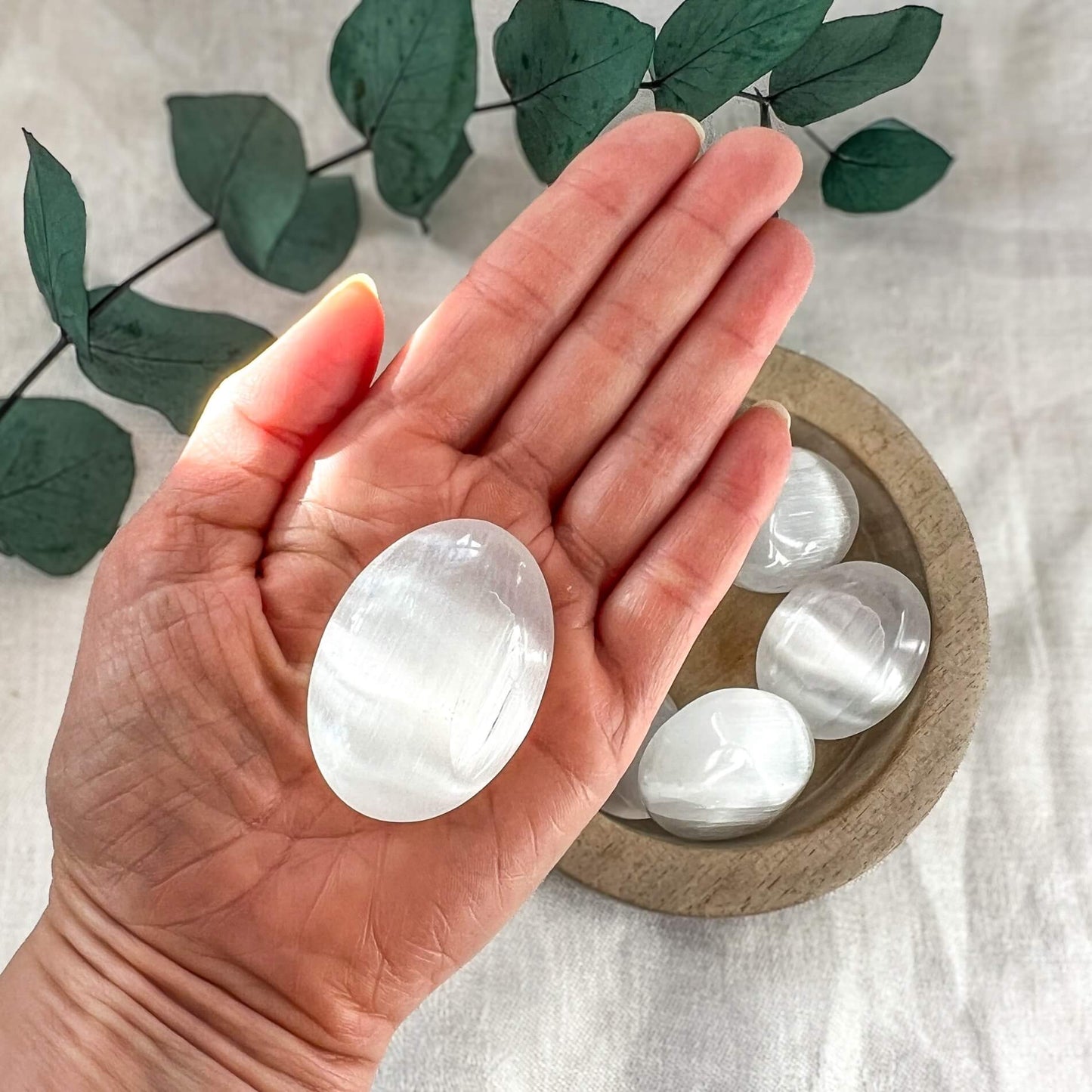 An open hand with a small oval white selenite crystal palmstone with a white backdrop, dish of selenite stones and eucalyptus leaves