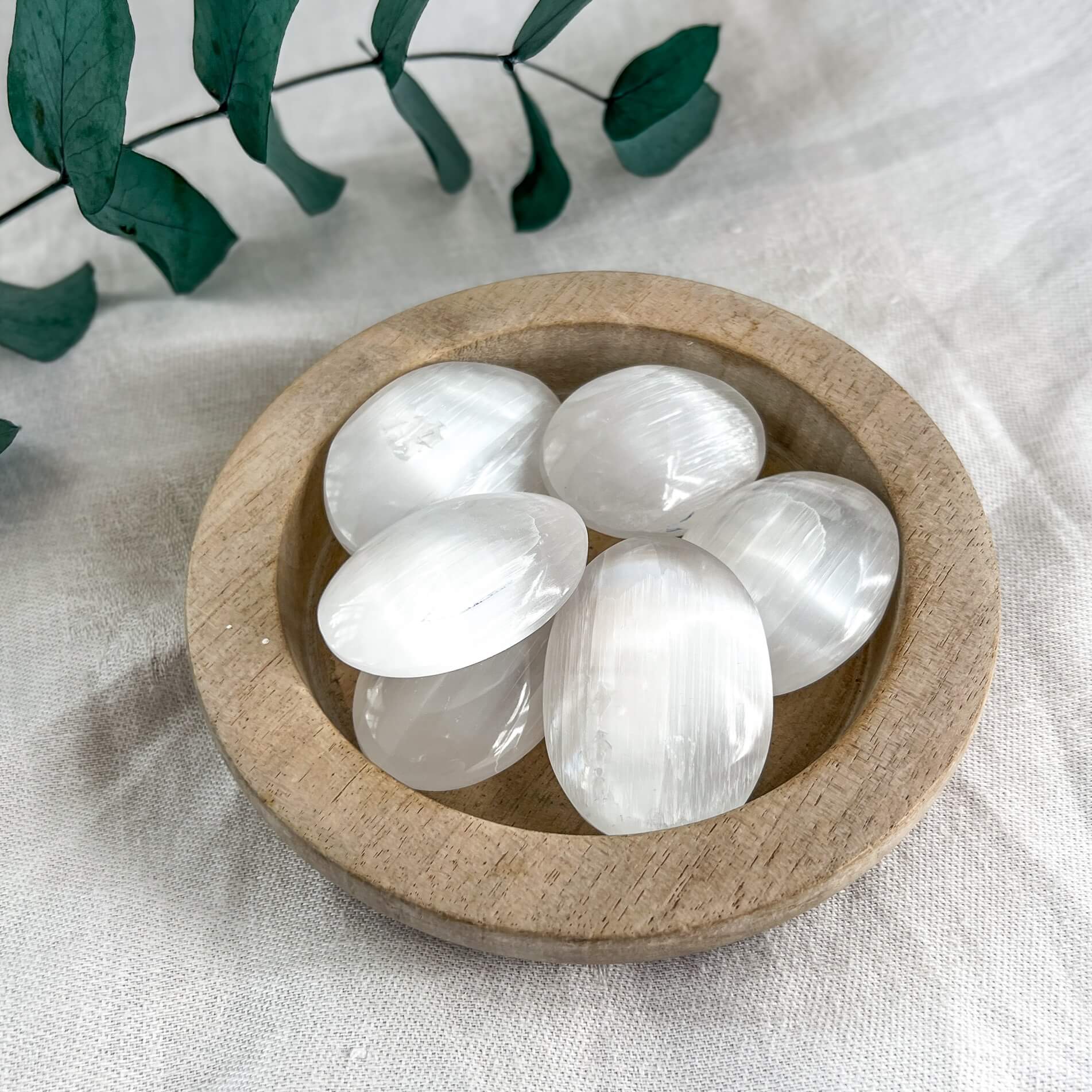 A small wooden dish of small oval white selenite crystal palmstones with a white tablecloth and eucalyptus leaves