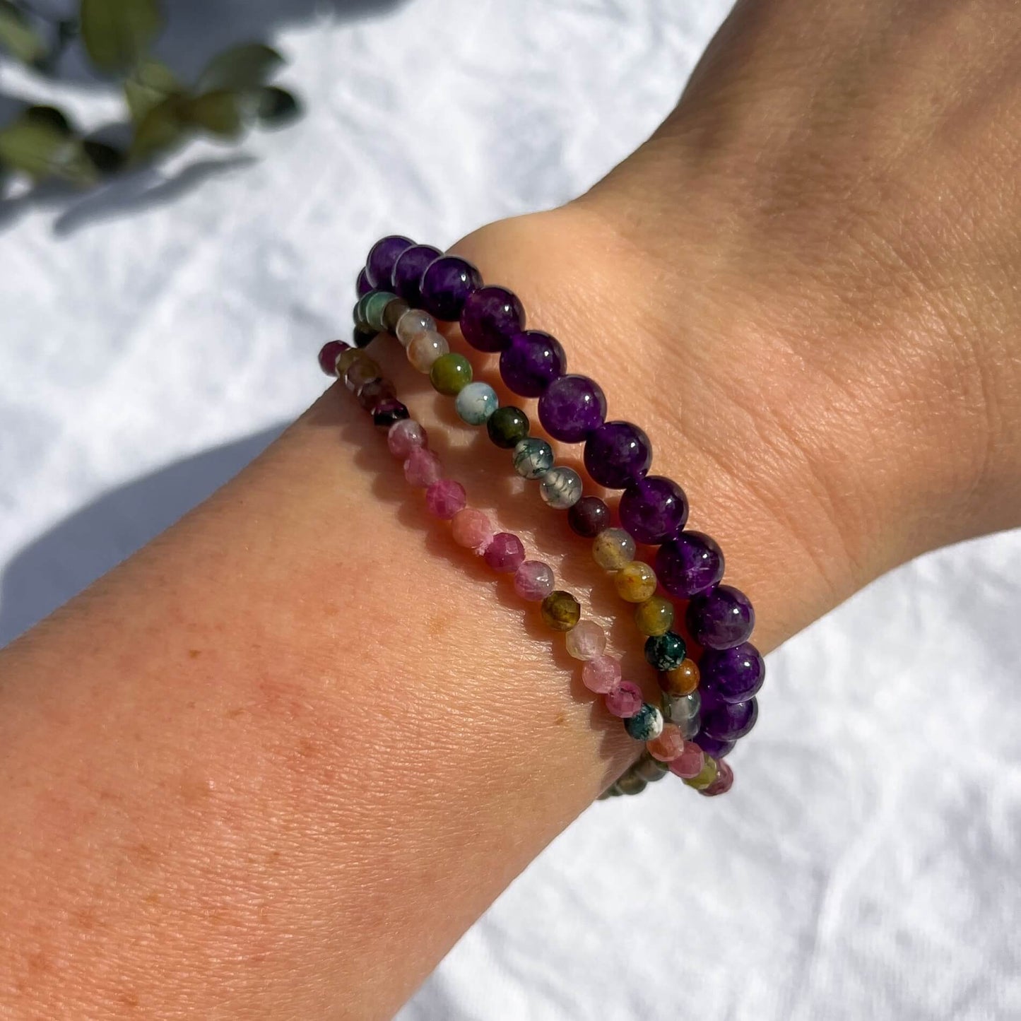 A woman's wrist wearing three pink, purple and green crystal bead bracelets in amethyst, tourmaline and jasper