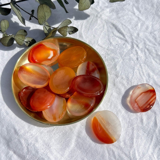 A brass dish filled with red and orange oval carnelian crystal thumb stones, two patterned carnelian crystal worry stones in the foreground