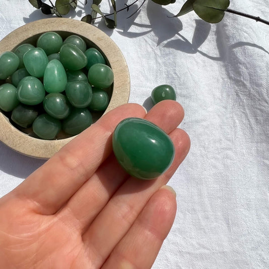 A green aventurine crystal tumblestone on an outstretched hand in the sun with a dish of green crystals in the background