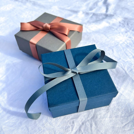 Luxury navy gift box tied in a teal grey ribbon with a grey gift box & pink ribbon in the background
