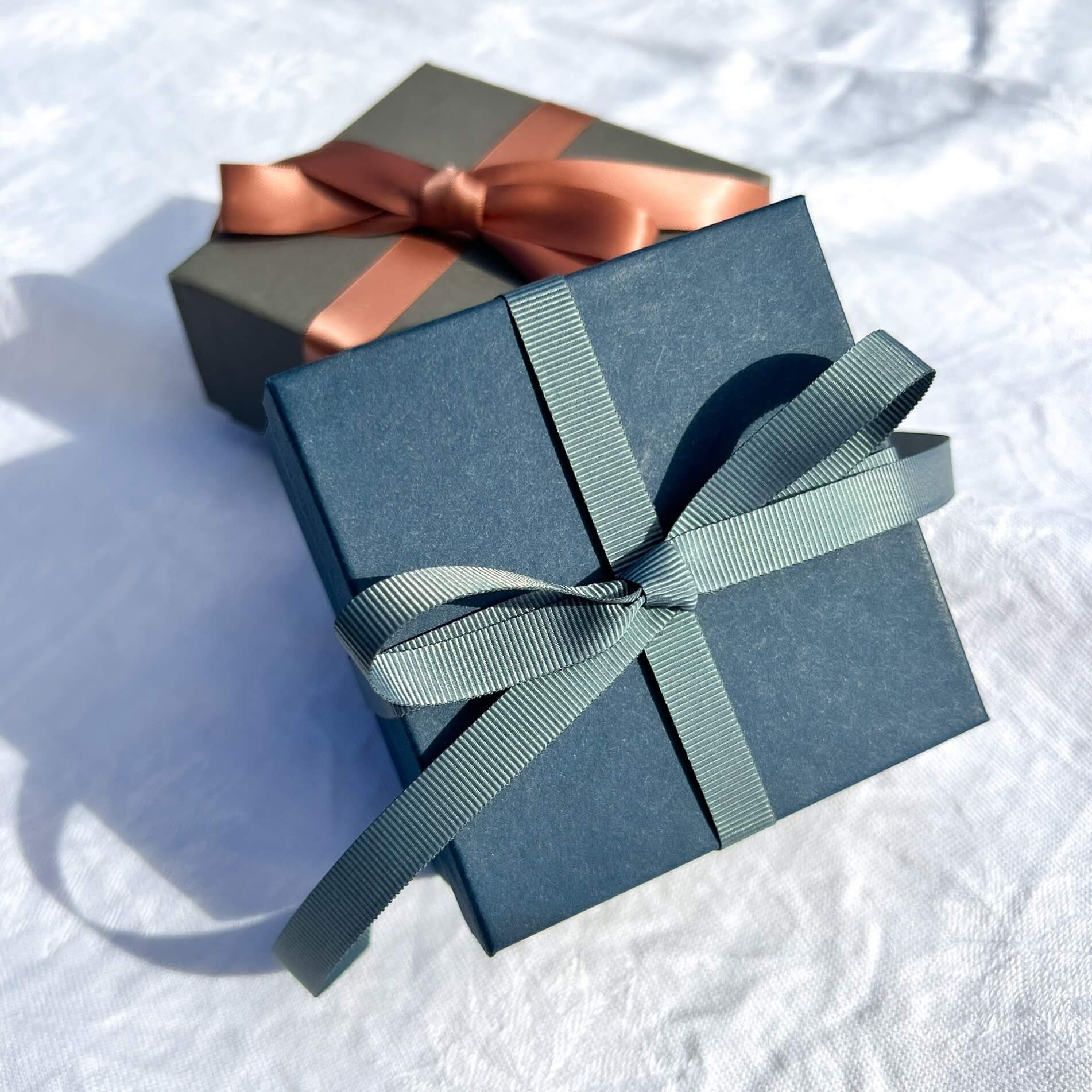 Luxury navy gift box tied in a teal grey ribbon with a grey gift box & pink ribbon in the background