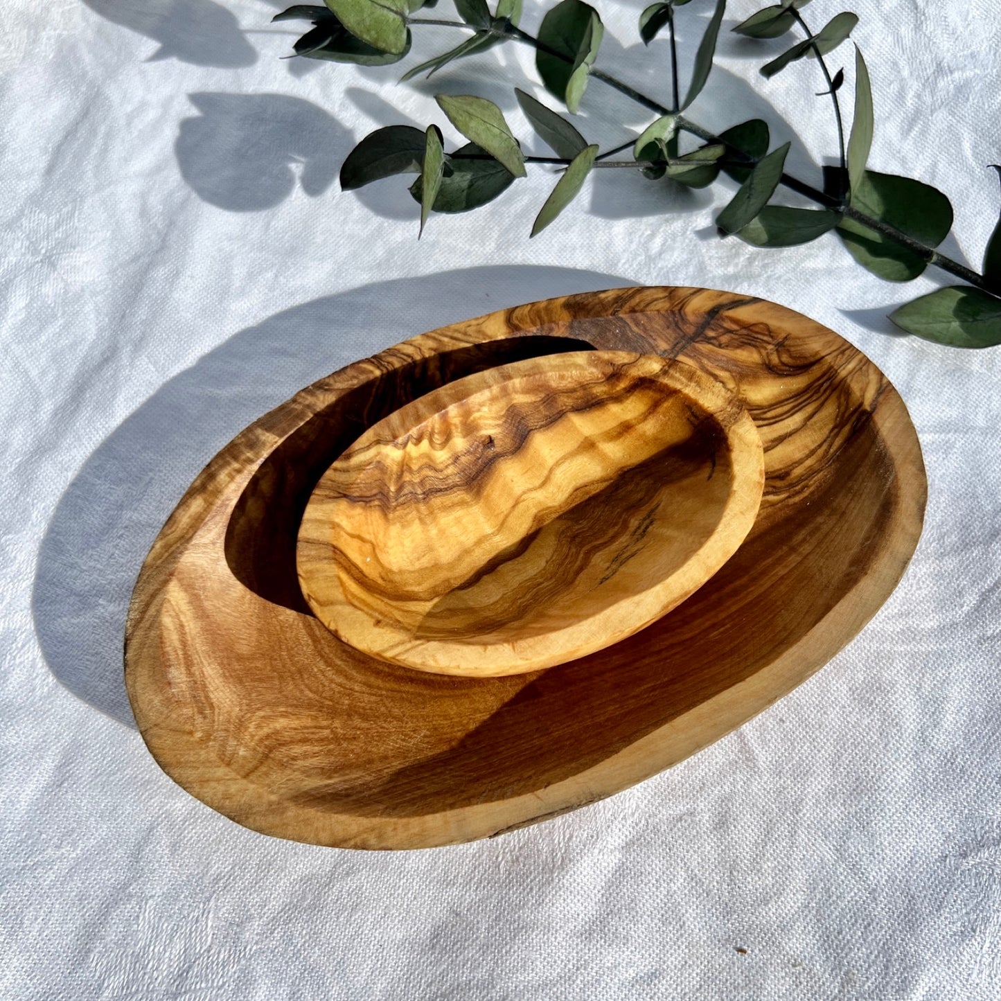 Two Warm honey coloured olive wooden bowls nested with eucalyptus leaves in the background